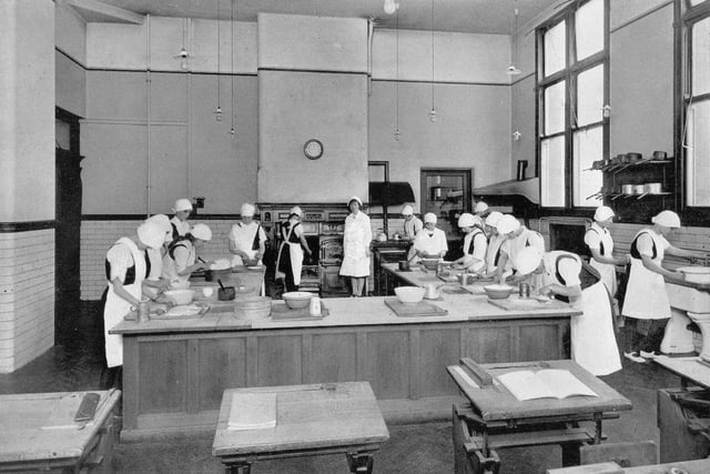 A cookery room in the girl's section of West Leeds High School. The pupils are dressed in white aprons and caps, and a range style cooker is visible at the back of the room.