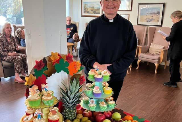Reverend from Blacklands Church stands beside a vibrant Harvest Festival display at Mountside Care Home, featuring fresh fruits and festive cupcakes for residents to enjoy