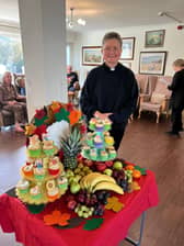 Reverend from Blacklands Church stands beside a vibrant Harvest Festival display at Mountside Care Home, featuring fresh fruits and festive cupcakes for residents to enjoy