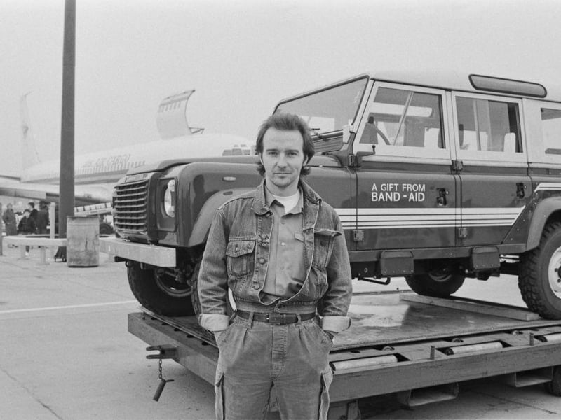 British singer-songwriter Midge Ure poses, hands in pockets, with a Land Rover Defender - 'A Gift From Band-Aid' written on the door - as the first air consignment of Band-Aid famine relief supplies to Ethiopia prepares to leave Gatwick Airport in Crawley on 9th March 1985.