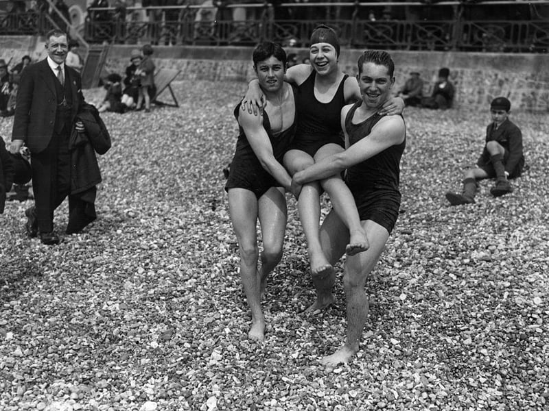 16th April 1927: Two young men helping a woman across the shingle on the beach at Brighton during the Easter Holiday.