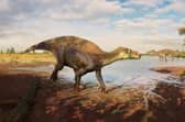 A reconstruction of the iguanodontian dinosaur Paludohadros proudlovii walking through mud along the edge of a shallow body of water. In the distance, the large predatory dinosaur Baryonyx can be seen. Picture courtesy of Guilherme Gehr/Natural History Museum