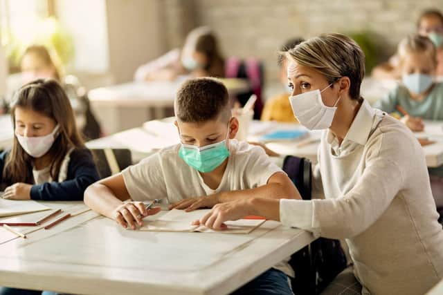 Elementary school teacher helping her student with learning during a class in the classroom. They are wearing protective face masks due to coronavirus pandemic. 