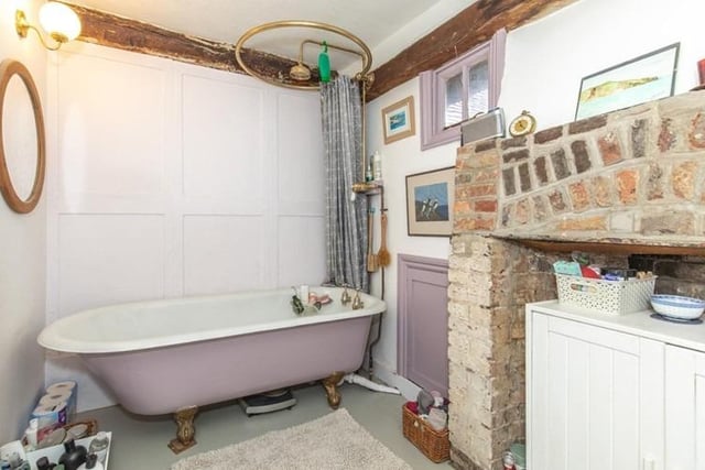 The family bathroom with a rubber tiled floor, roll top free standing bath with shower over, feature brick fireplace, wooden vanity unit with inset hand wash basin, wood panelled wall.